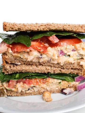 Two halves of a sandwich stacked on top of one another served on a white plate. The sandwich is filled with chickpea tuna salad, sliced tomatoes, red onion, and spinach.