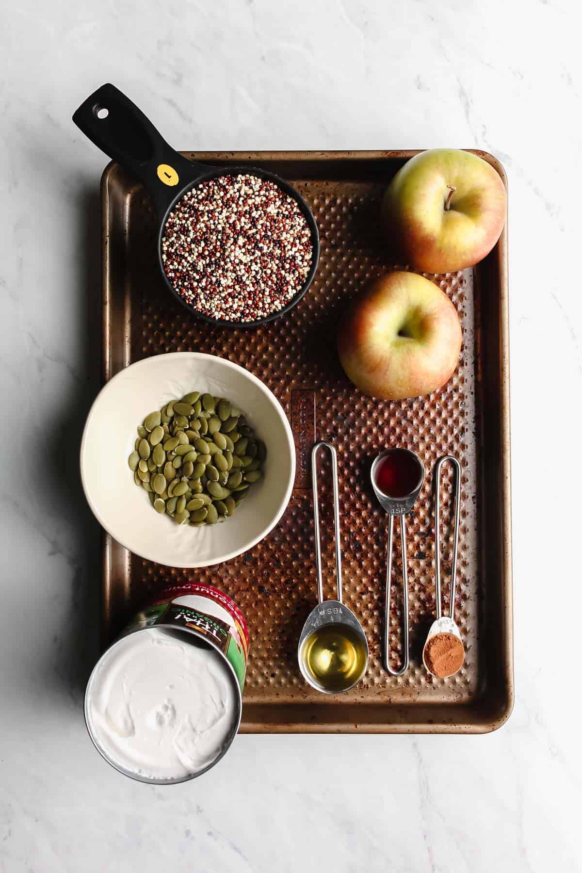 Overhead view of all of the ingredients on a baking sheet which is sitting on a marble background. 2 small apples, uncooked quinoa in a black measuring cup, raw pepitas in white bowl, opened can of coconut milk, 3 measuring spoons that contain cinnamon, avocado other oil, and vanilla extract.
