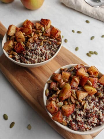 Coconut milk breakfast quinoa topped with sautéed apples and pepitas served in a cream colored bowl. Both bowls are sitting on a wooden platter and there are some loose peptitas spilled out next to the wooden platter. There are also two silver spoons sitting on a folded beige cloth napkin.