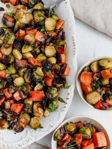 Roasted brussels sprouts, sweet potato, and red onion served on a white serving plater and well as served in 2 cream colored bowls.