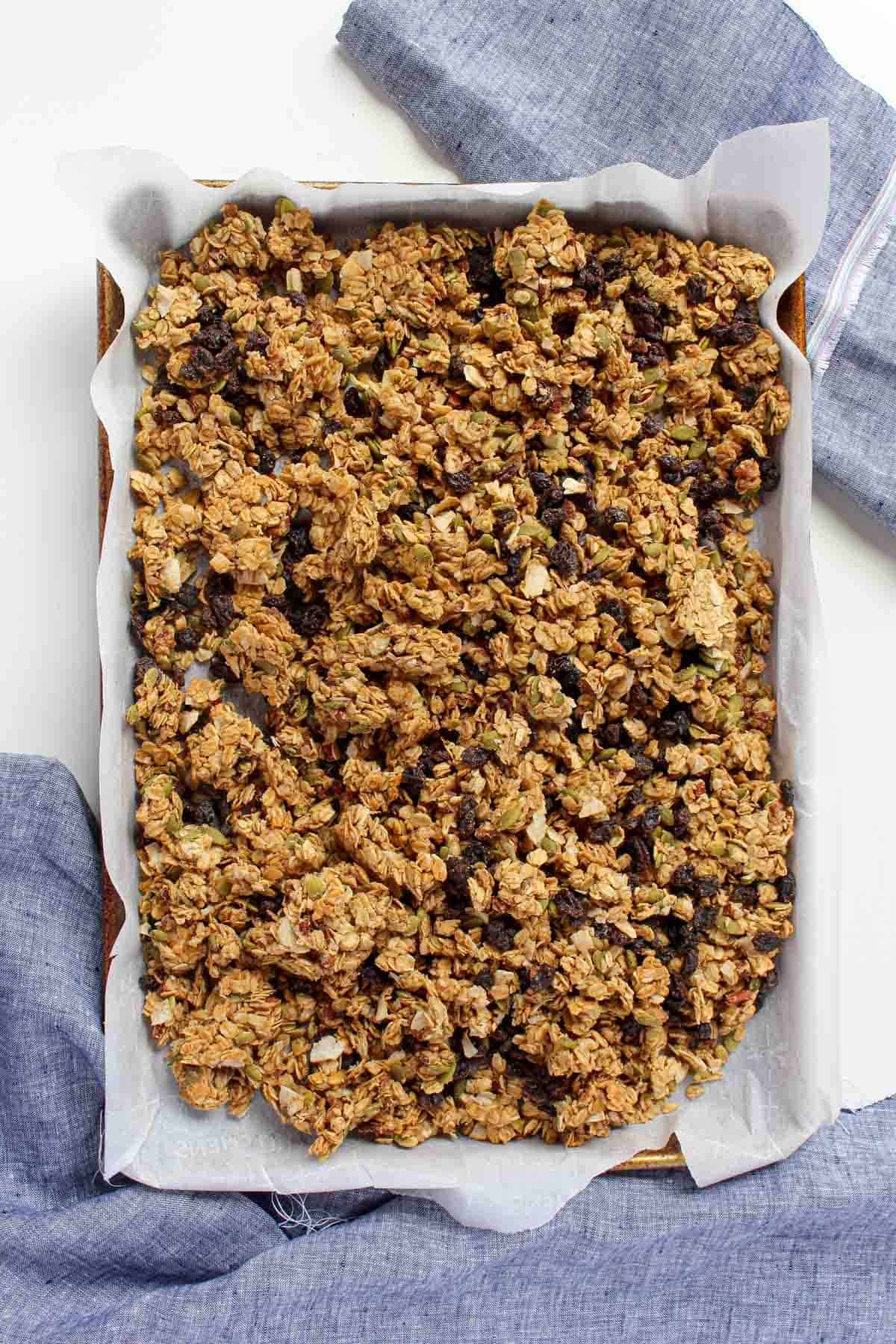 Homemade peanut butter granola on baking sheet lined with white parchment paper with two blue napkins in the background.