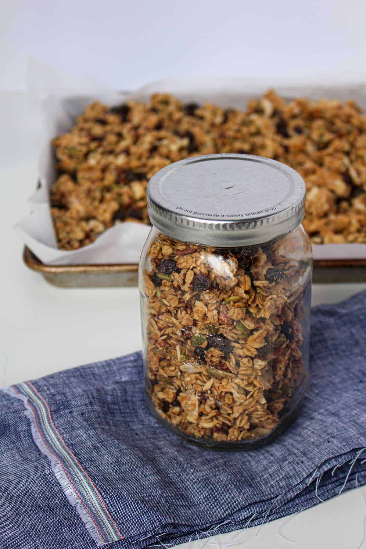 Mason jar filled with peanut butter granola on top of a blue napkin with a baking sheet lined with white parchment paper full of granola in the background.