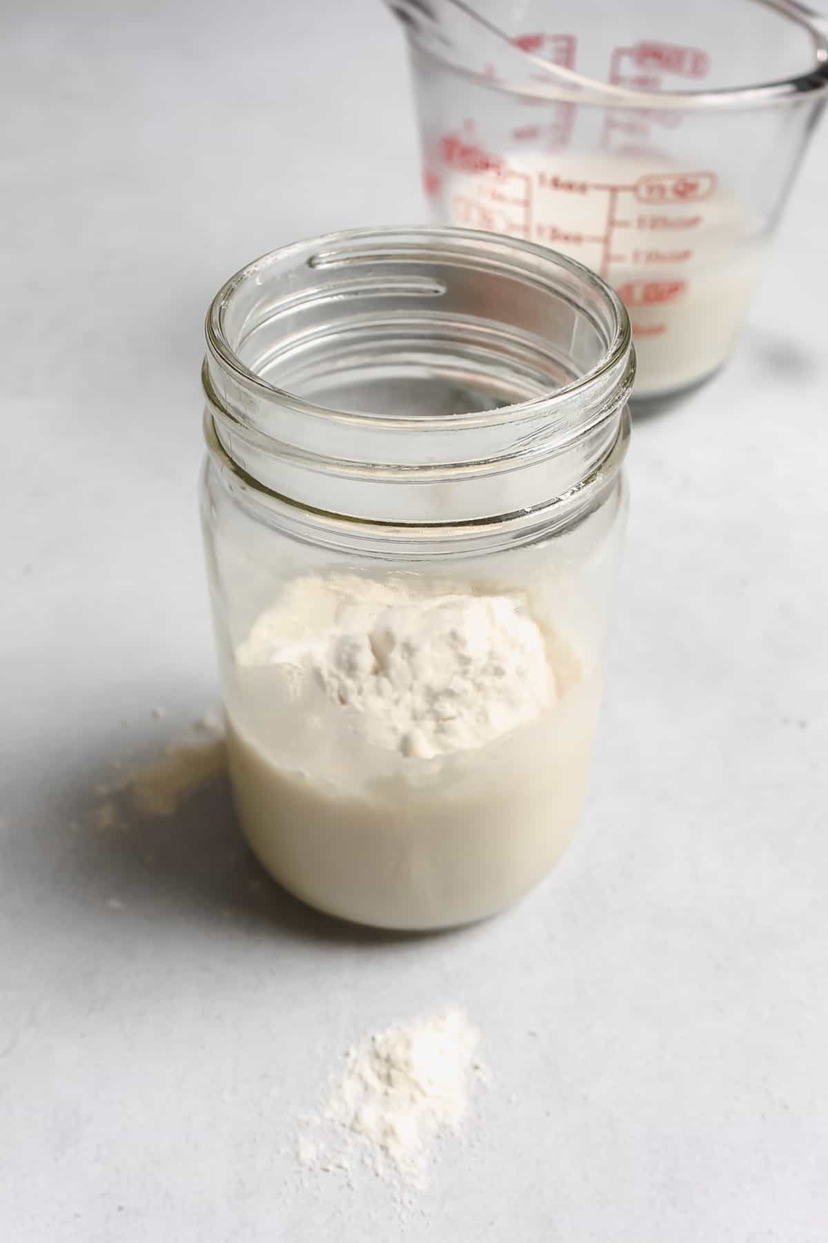 Small glass jar filled with vegan half and half and white flour. In the background there is a measuring cup also filled with vegan half and half.