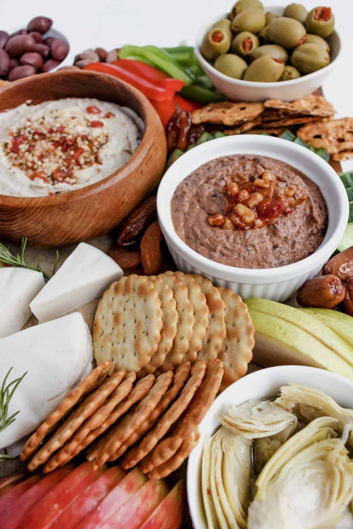 Closer view of bean dip in a ramekin, hummus in a round wood bowl, green olives, crackers, and marinated artichokes