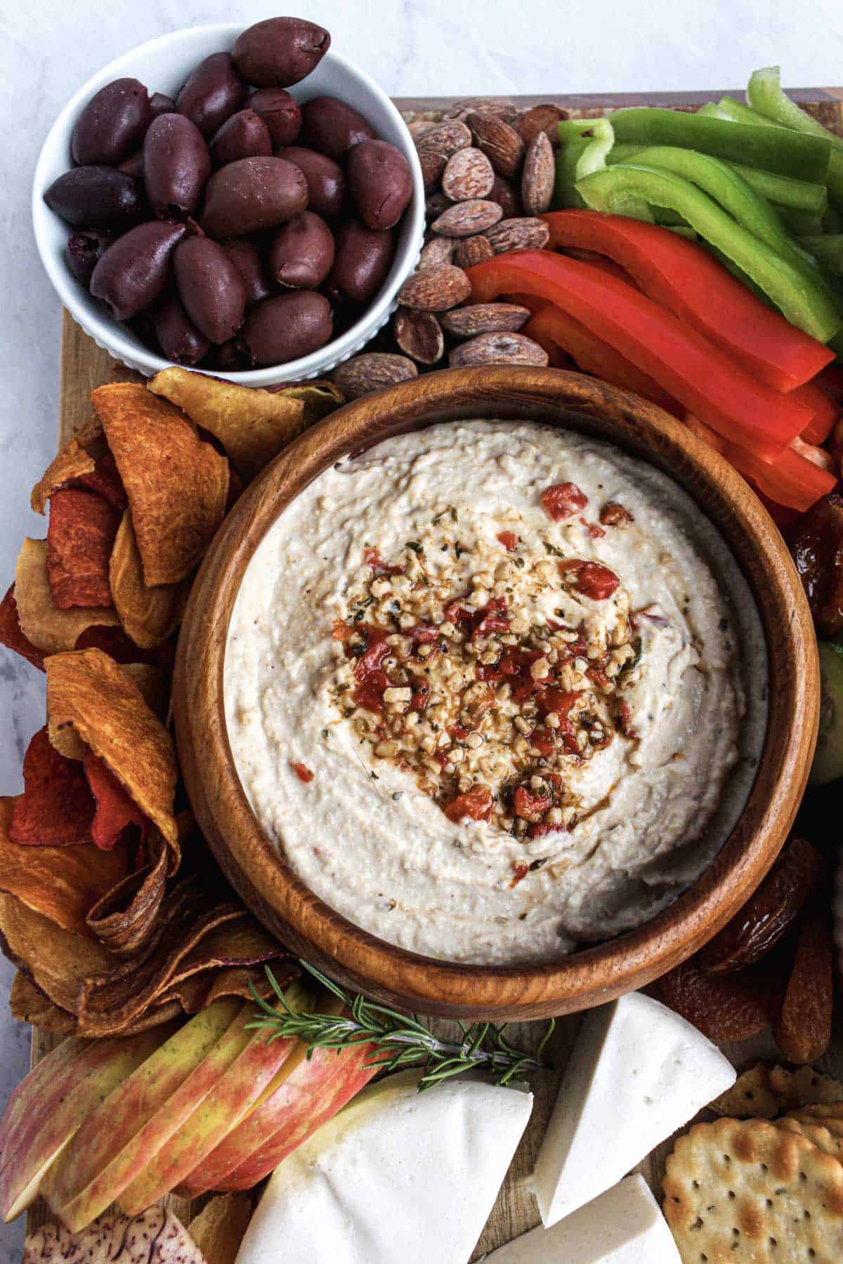 Kalamata olives in a smaller white bowl, salted almonds, sliced green and red bell pepper, hummus in a wooden bowl, veggie chips, sliced red apple, part of vegan cheese wheel.