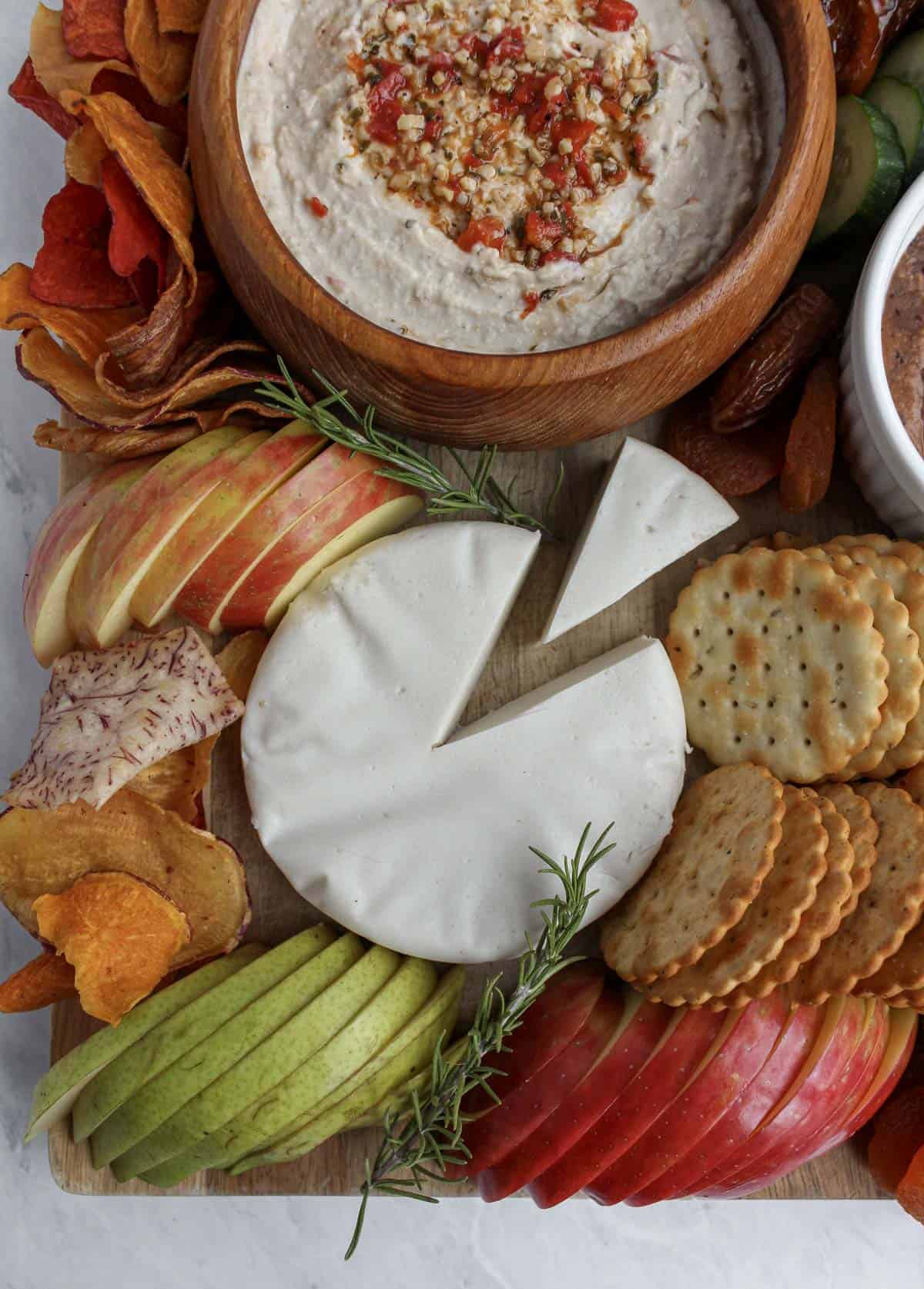 Hummus in medium wooden bowl, sliced red apple, sliced green pear, crackers, veggies chips, a white wheel of vegan cheese with a triangle cut out of it, and a sprig of rosemary for garnish.