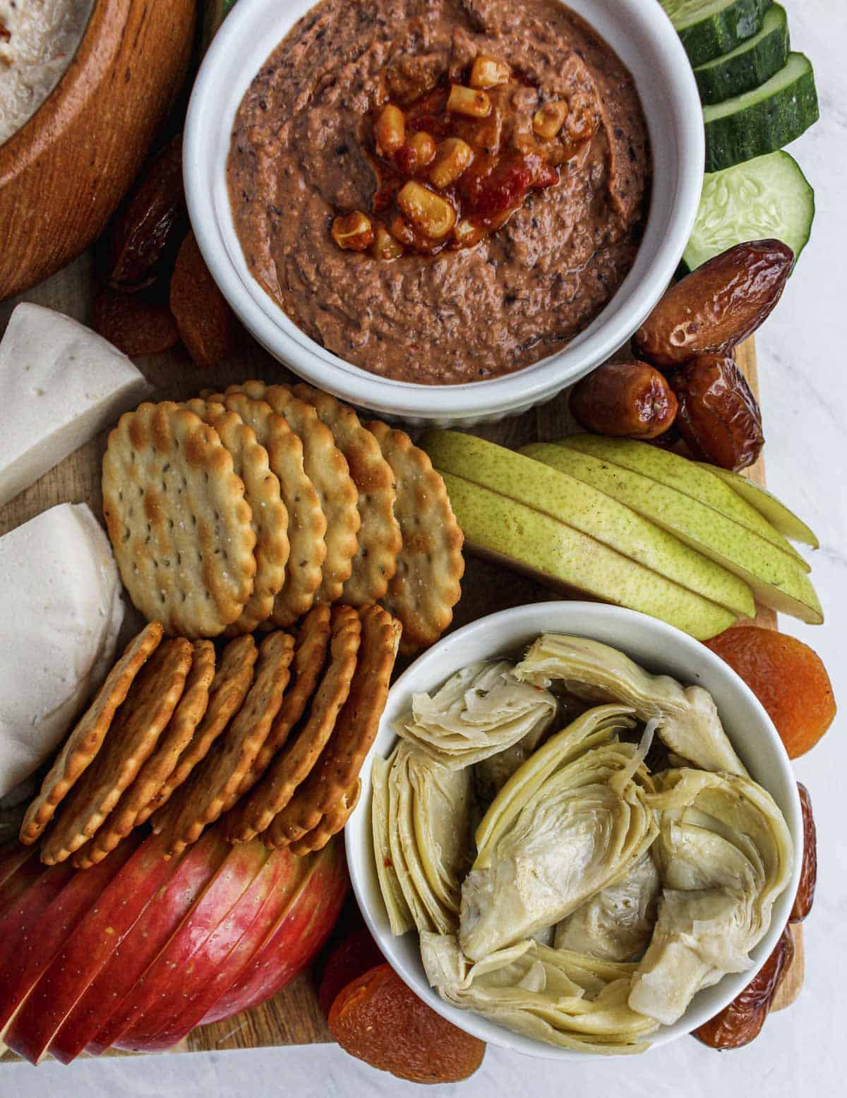 Marinated artichokes in a small white bowl, dried apricots, sliced red apple, crackers, sliced green pear, sliced cucumber, bean dip in a smaller white ramekin.