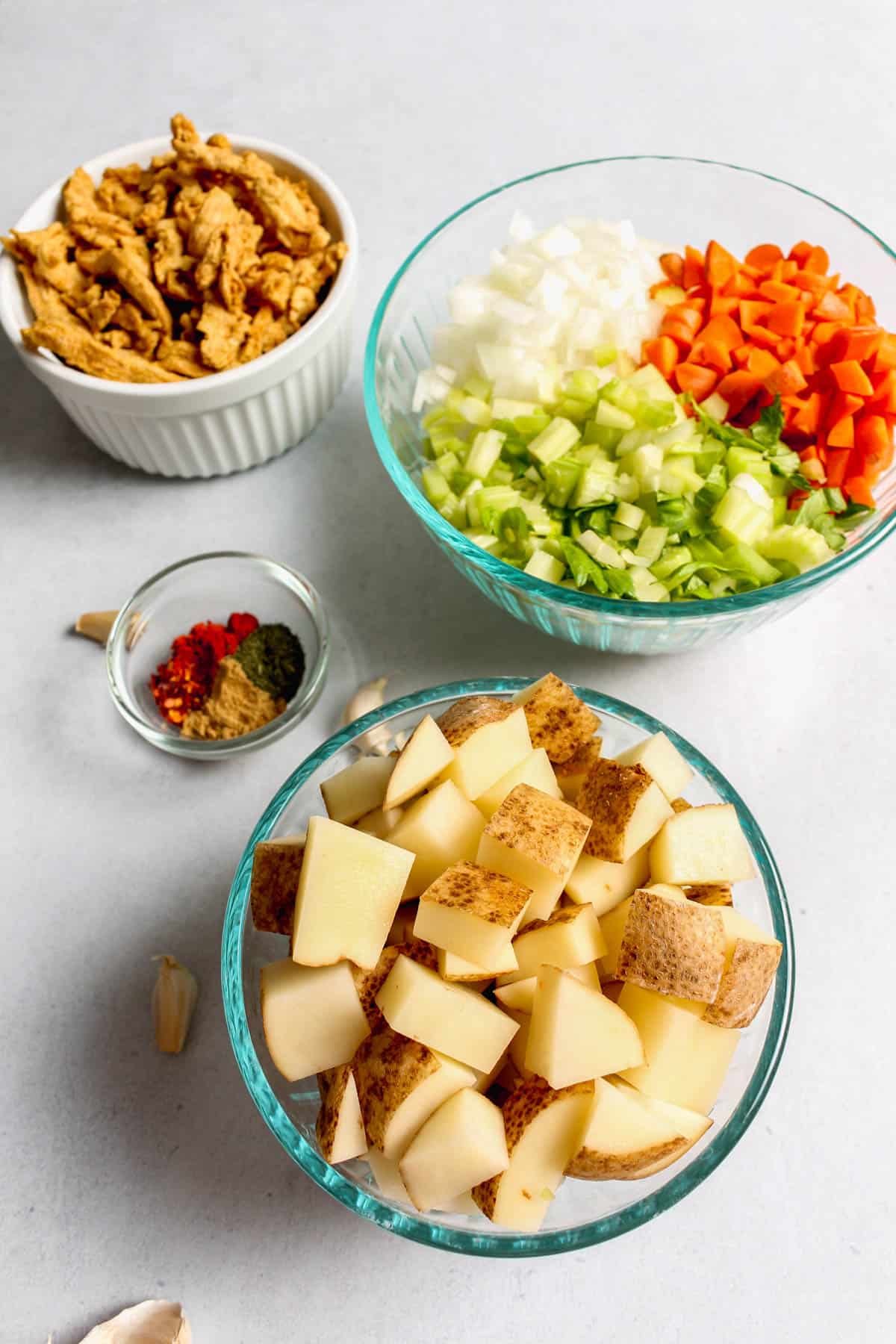 Ingredients for the soup, washed and prepared and separated in different bowls: cubed potatoes in a glass bowl, chopped carrots, onion and celery together in a glass bowl, dried soy curls in a white bowl, spices in small glass prep bowl, and few loose garlic cloves on a grey background