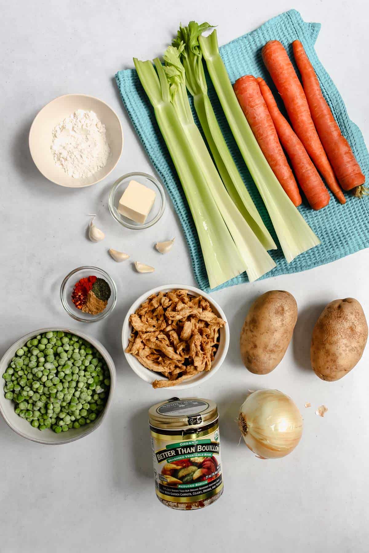 All ingredients for soup on a grey background. There are 4 celery stalks and 4 large carrots on a blue towel, a yellow onion, 2 potatoes, jar of Better Than Bouillion Base, Soy curls, frozen peas, spices, garlic cloves, vegan butter, and flour.