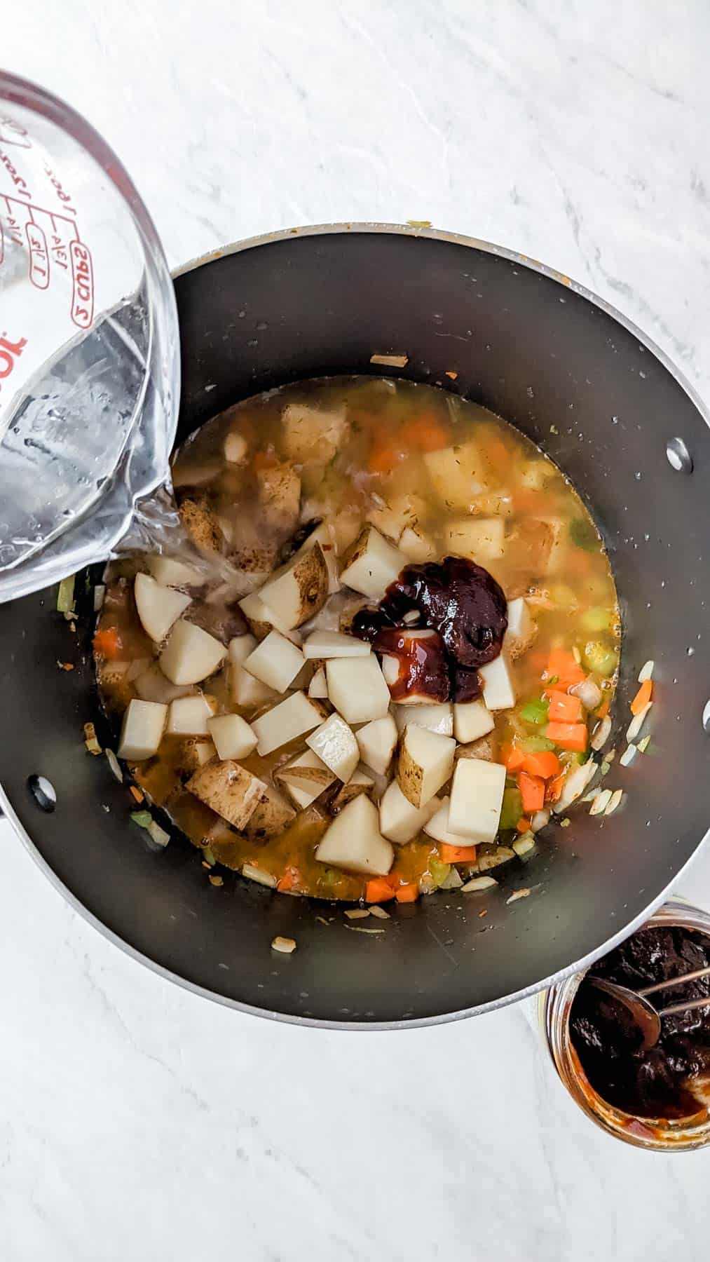 Water being poured in the soup from a glass measuring cup. Soup pot with potatoes and Better Than Bouillon Base on top of the other ingredients in the pot. A open bar of Better Than Bouillon Base with a silver measuring spoon in it.