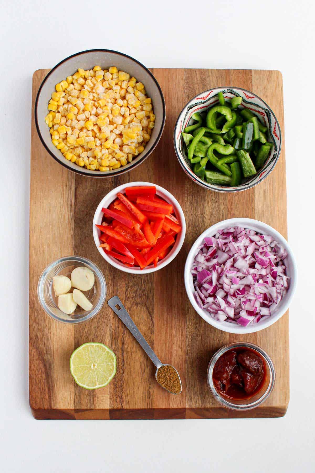 Individual bowls filled with frozen corn, sliced green and red bell peppers, chopped red onion, half of a lime, fresh garlic, 1 teaspoon filled with cumin, and chipotle peppers in sauce
