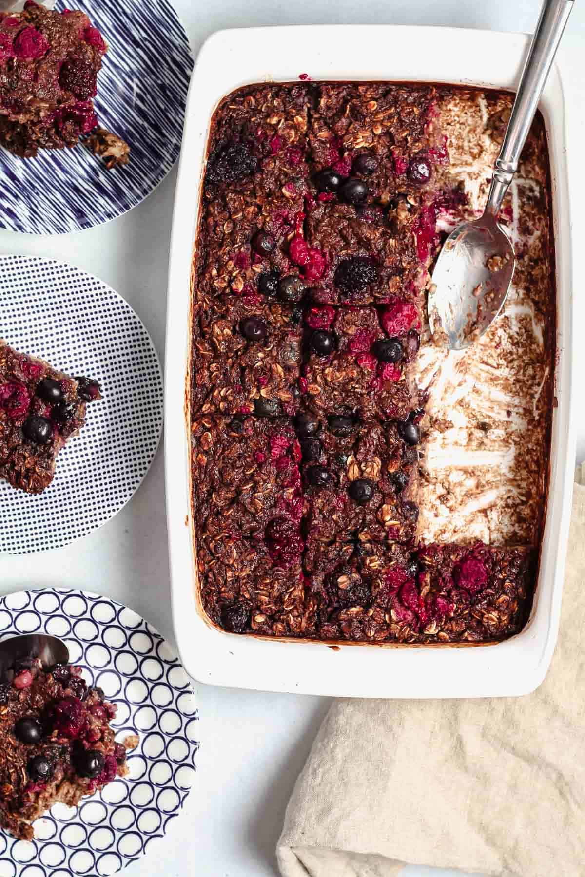 Three pieces of chocolate banana baked oatmeal served on three blue and white plates along side a white baking dish holding the rest of the baked oatmeal with a silver spoon along side a tan cloth napkin.