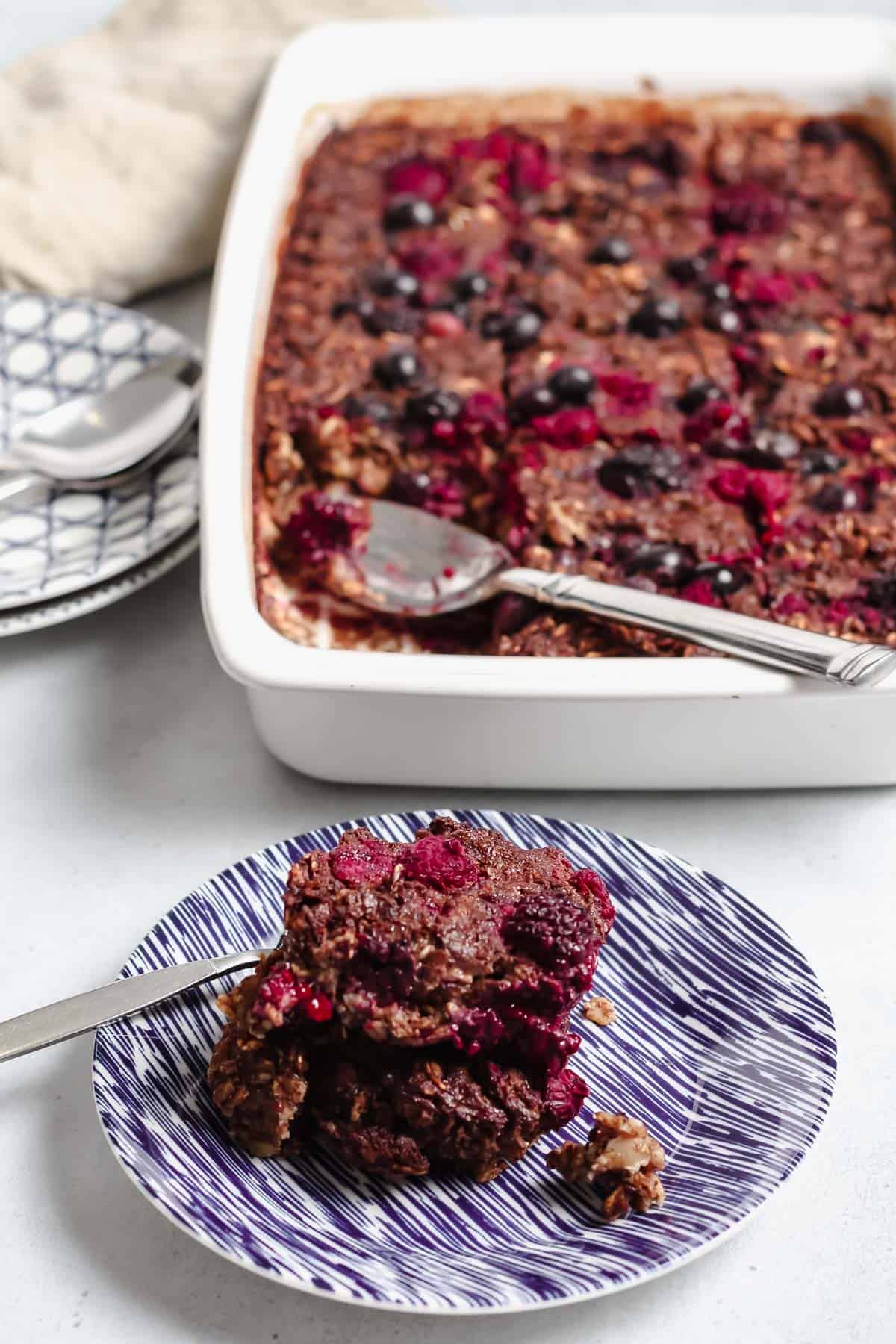 Serving of baked oatmeal on a blue and white pattered plate. White baking dish holding the rest of baked oatmeal in the background.