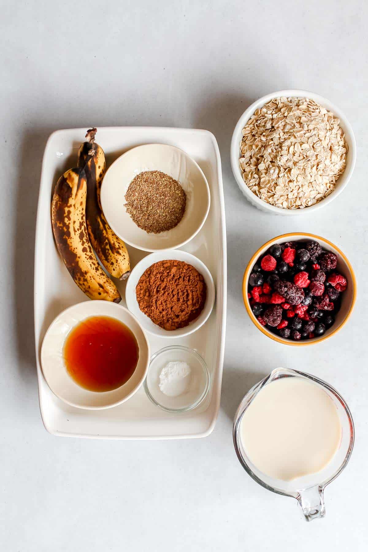 Ingredients for chocolate banana baked oatmeal with mixed berries in measuring cups on white background