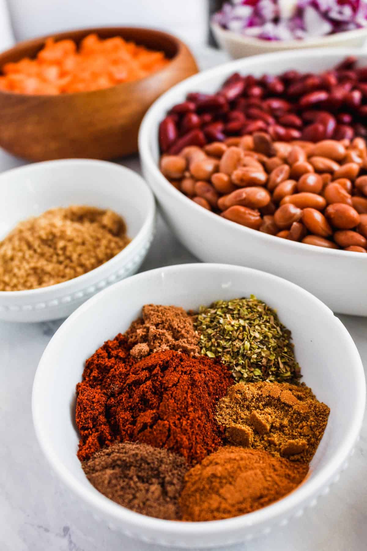 Close up of spices used for chili in small white bowl, bowls in the background with beans, brown sugar, carrots, and red onion.