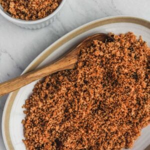 Cooked garlic chipotle quinoa on white serving platter with a wooden spoon on a marble background. Bowl of quinoa in upper lefthand corner.