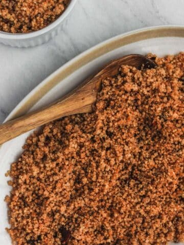 Cooked garlic chipotle quinoa on white serving platter with a wooden spoon on a marble background. Bowl of quinoa in upper lefthand corner.
