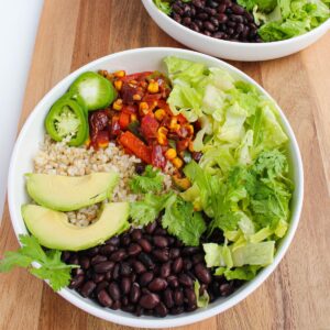 White bowl sitting on a wooden cutting board filled with black beans, cooked peppers, onions, and corn, fresh lettuce, slices of avocado and jalapeño