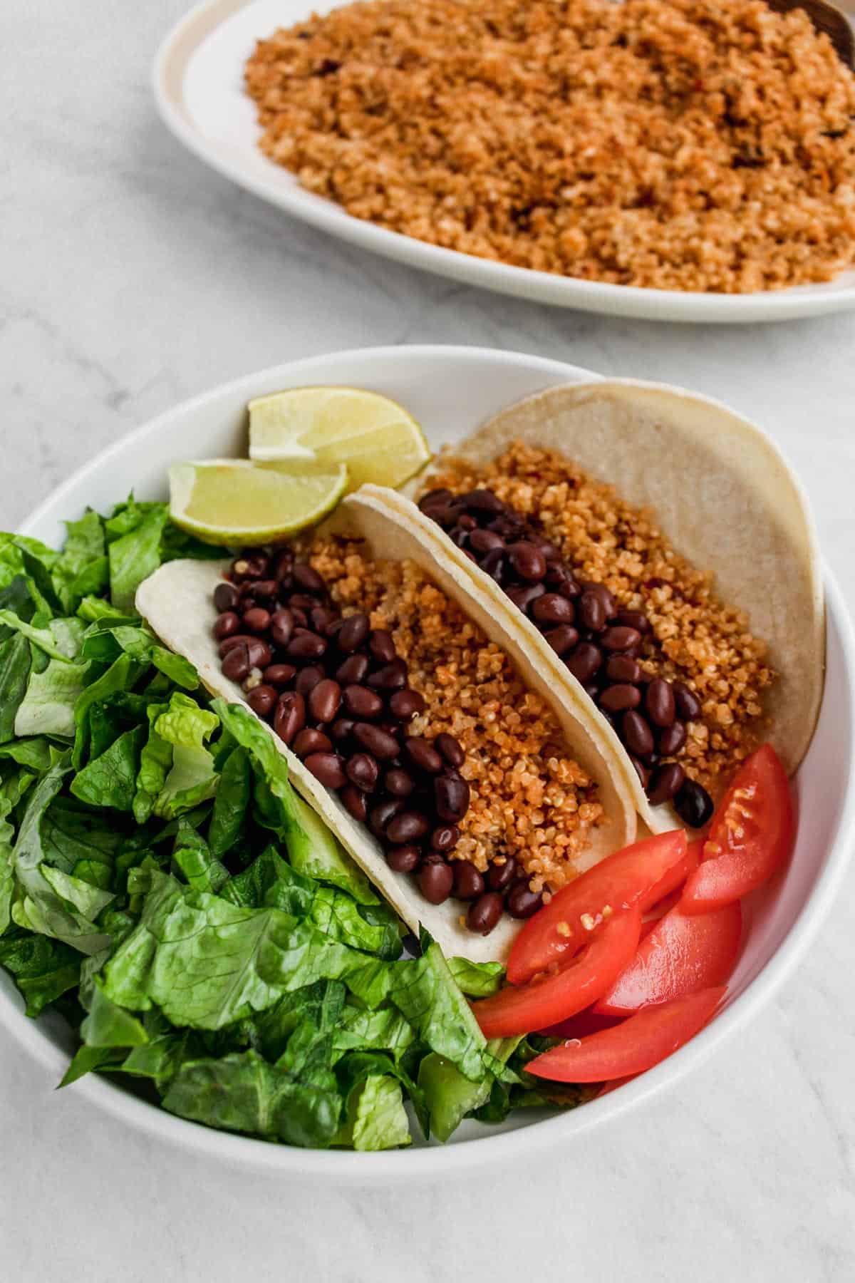 Two tacos filled with black beans and quinoa, with lettuce, tomato and two slices of lime on the side. Serving platter of quinoa in the background.