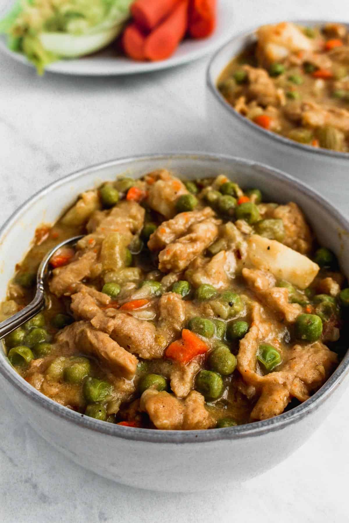 Two grey bowls with vegan chicken and potato soup and a white plated in the background with celery and carrots on it.