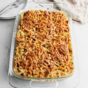 Cooked vegan chicken casserole in a glass dish on white back ground along with two stacked white plates with two forks and a tan napkin.