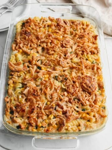 Cooked vegan chicken casserole in a glass dish on white back ground along with two stacked white plates with two forks and a tan napkin.