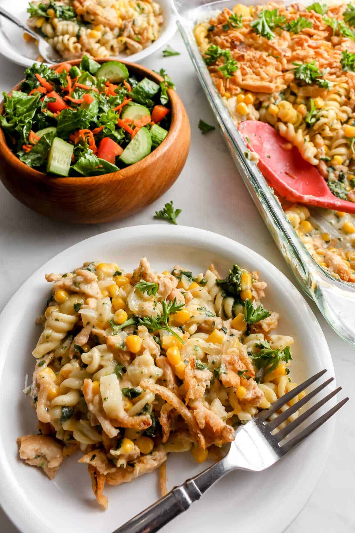 Cooked casserole plated served on white plates with silver forks. Salad served in a wooden bowl. Casserole dish with red serving spoon off to the right side.