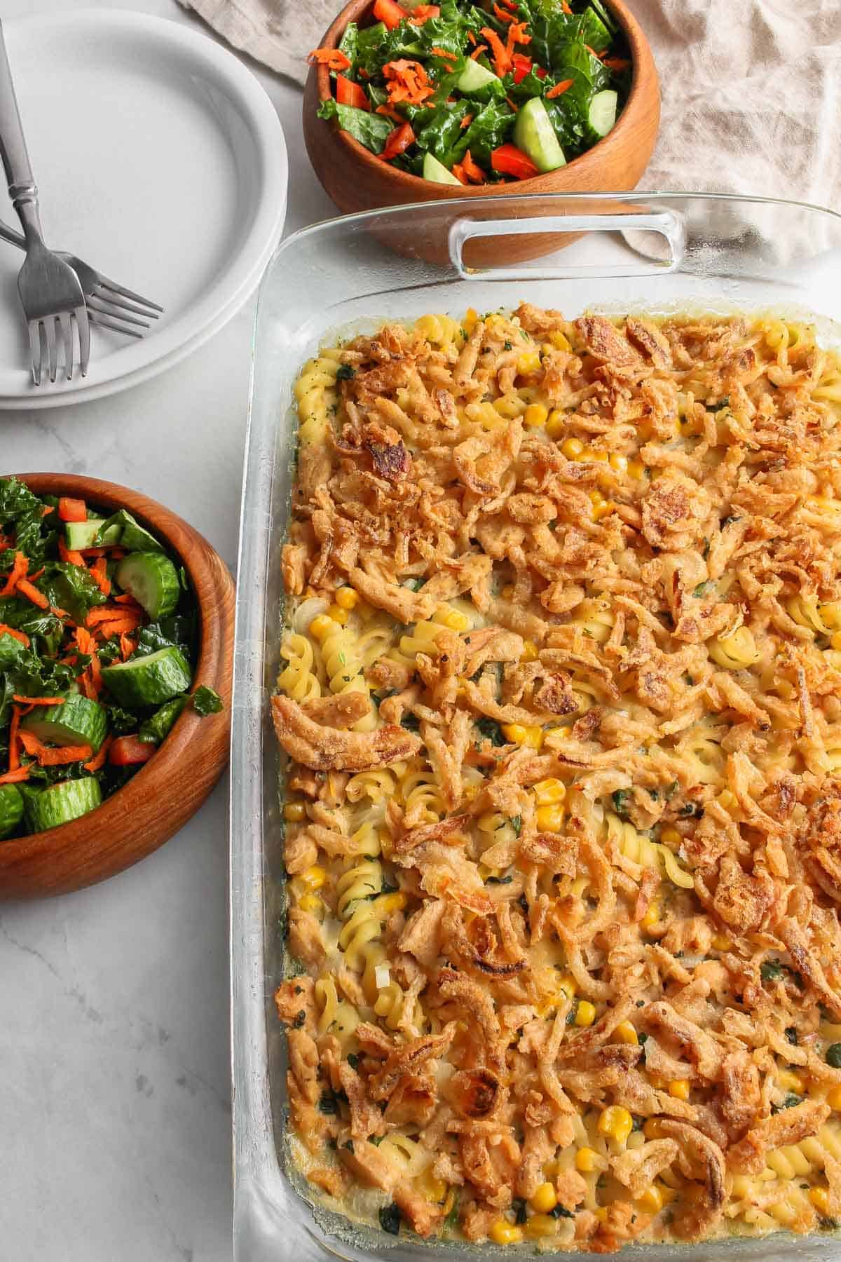 Cooked casserole in glass baking dish with two bowls of salad and two white serving plates with two forks on a white, marble background.