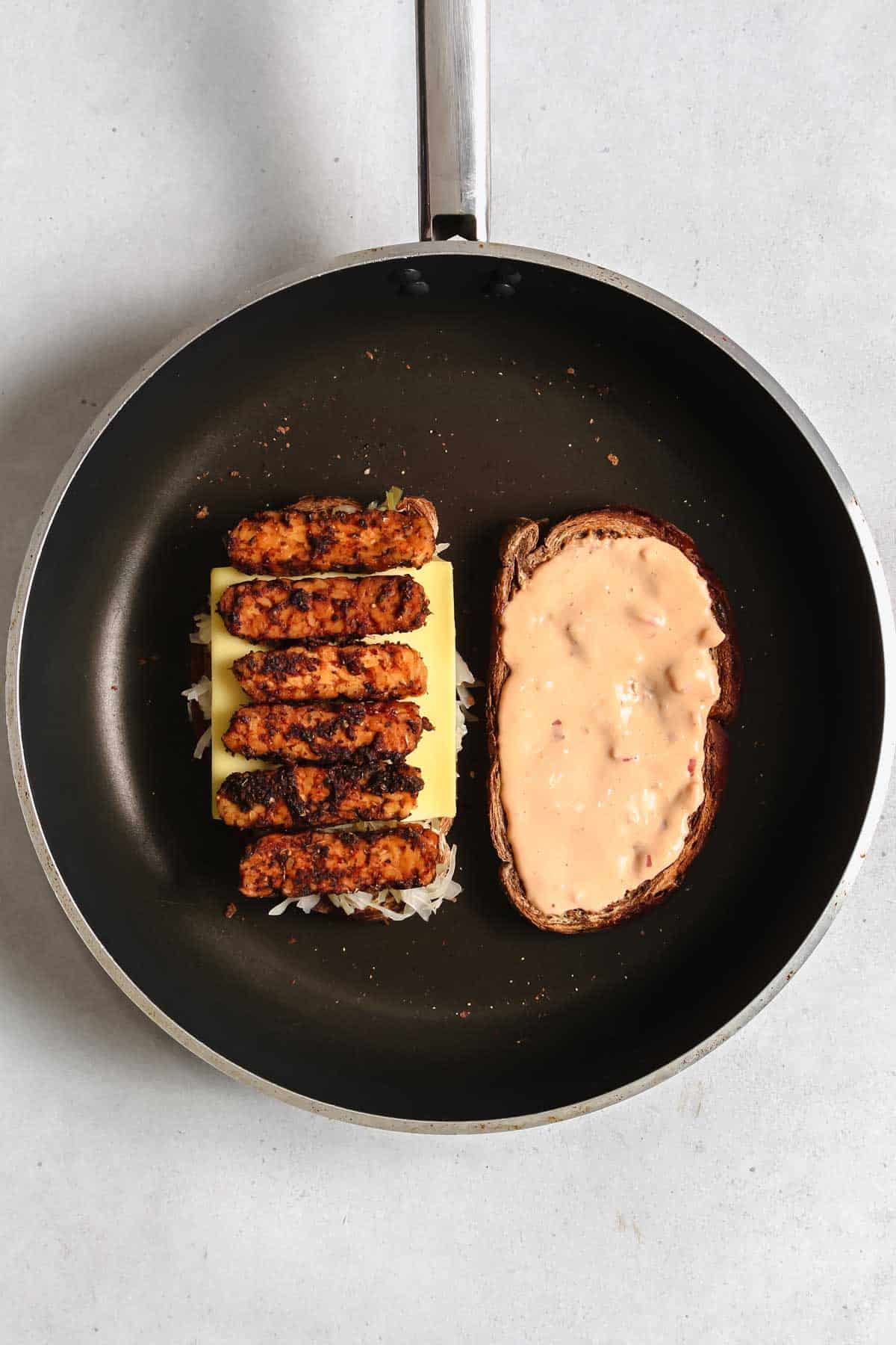 Two slices of bread in large pan: one with with tempeh, sauerkraut, sliced vegan cheese and the other with dressing on it.
