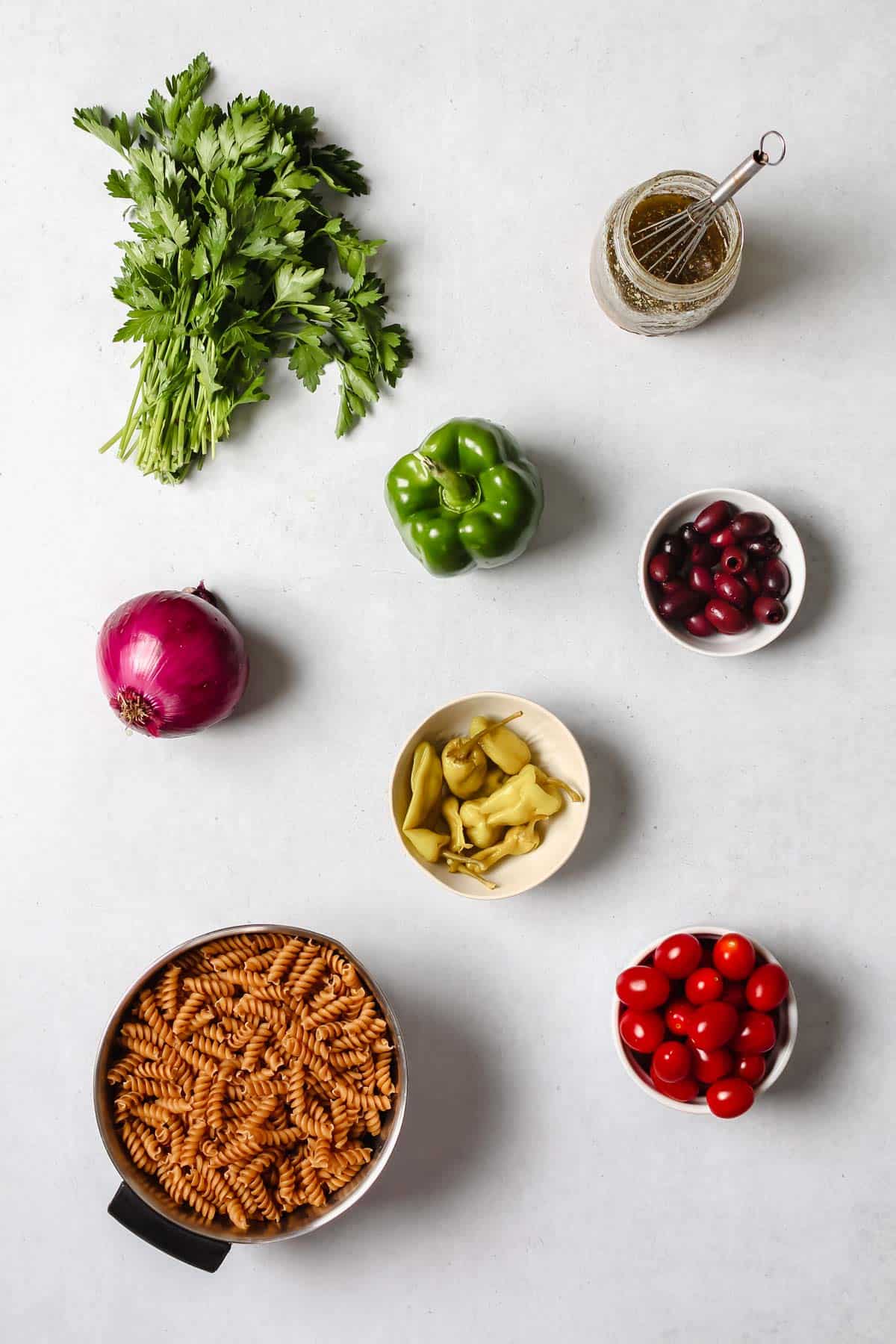 Individual pasta ingredients: fresh parsley, glass jar of dressing, green bell pepper, red onion, Kalamata olives, pepperoncini peppers, grape tomatoes and uncooked pasta on white background