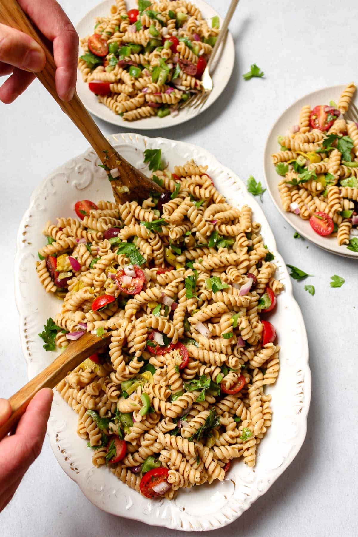 Vegan pasta salad on a white platter being served with two wooden serving spoons with two plates of pasta salad on the side.
