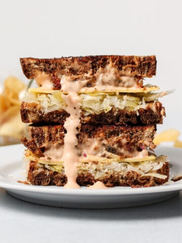 Two halves of a vegan tempeh reuben stacked on top of each other on a white plate.