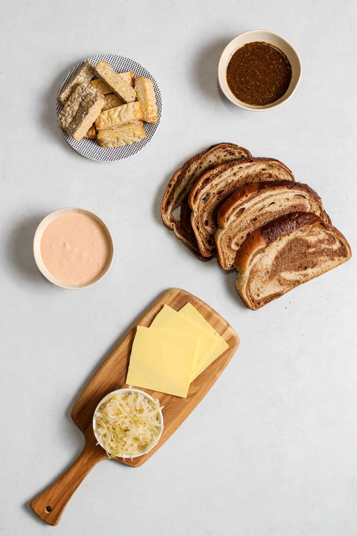 Individual ingredients for reuben: sliced tempeh on blue plate, marinate and Russian dressing in bowls, 4 slices of bread, and sliced cheese and a small bowl of sauerkraut on wooden platter.