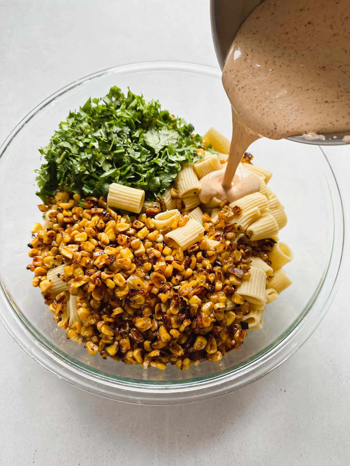 Large glass mixing bowl filled with charred corn, cooked pasta, and cilantro with dressing being poured on from above from a stainless steel bowl.