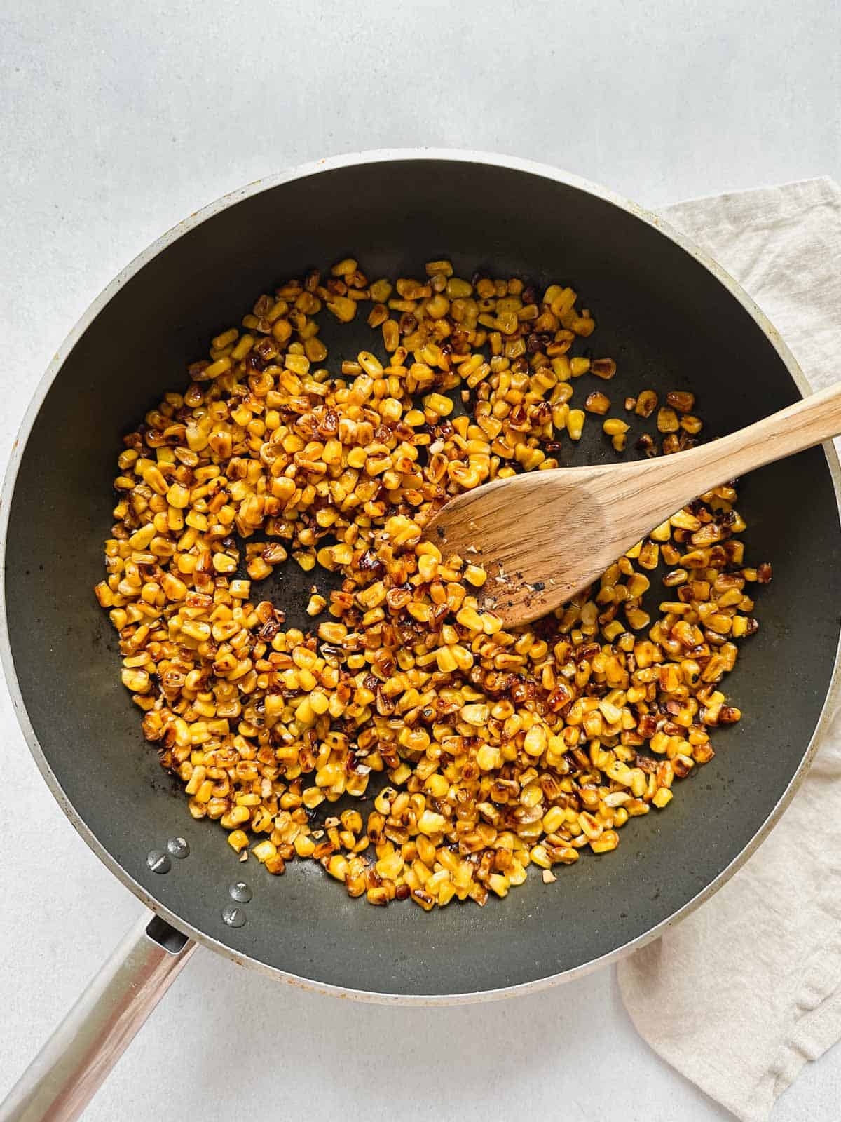 Large skillet filled with charred corn and a wooden spoon on a white background with a tan napkin on the right side.