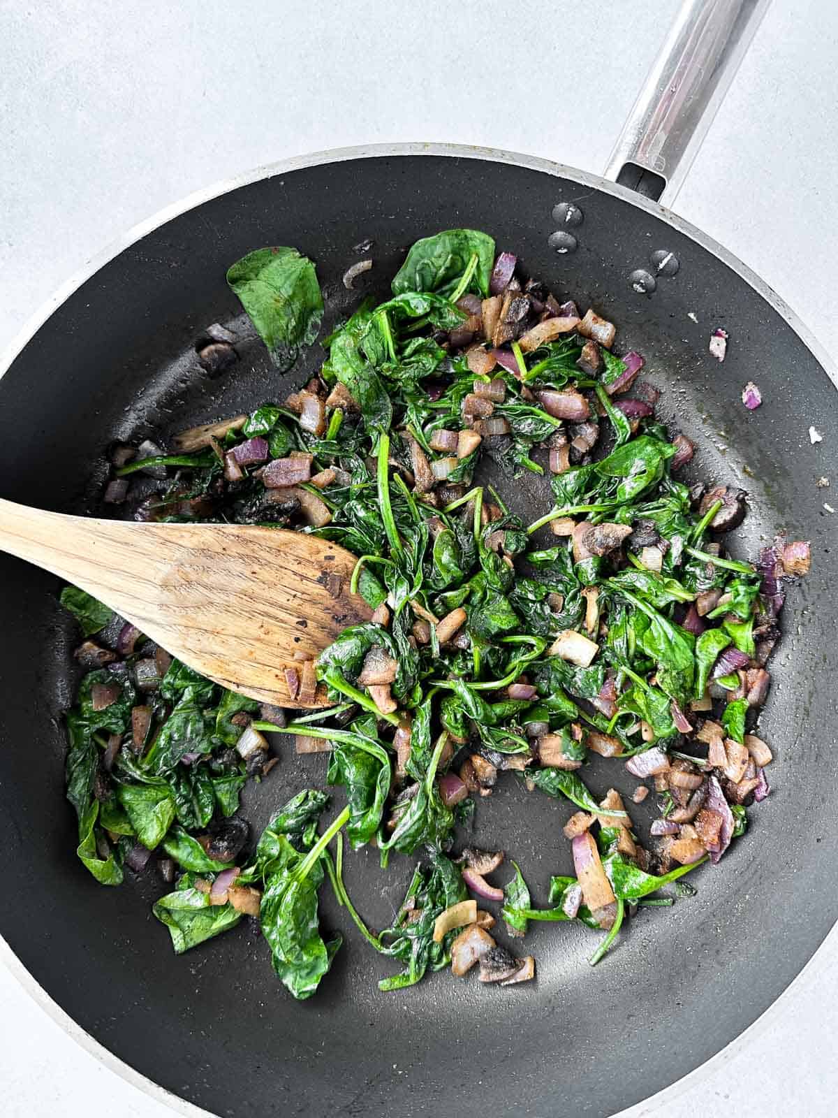 Sautéed spinach, mushroom and red onion in black skillet with wooden spoon on white background