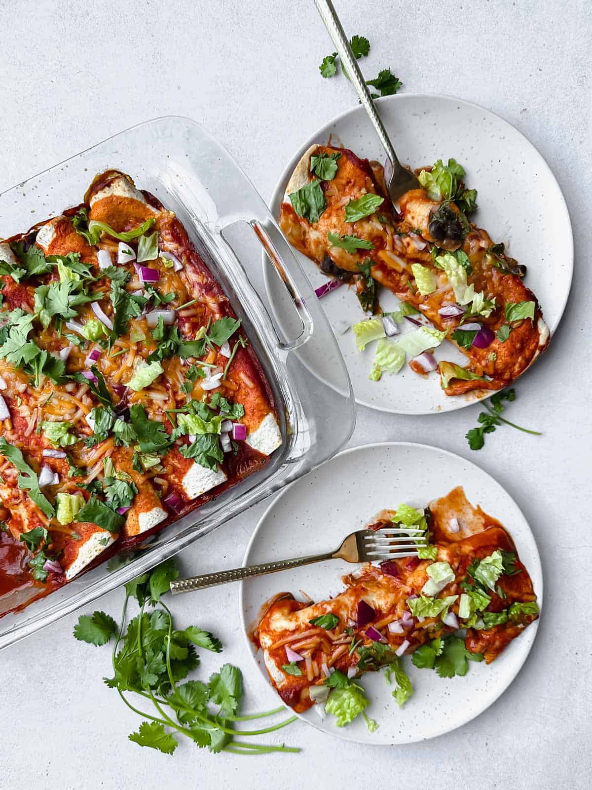 Two plated vegan black bean enchiladas on white plates with forks, glass baking dish with cooked enchiladas topped with lettuce, cilantro and red onion
