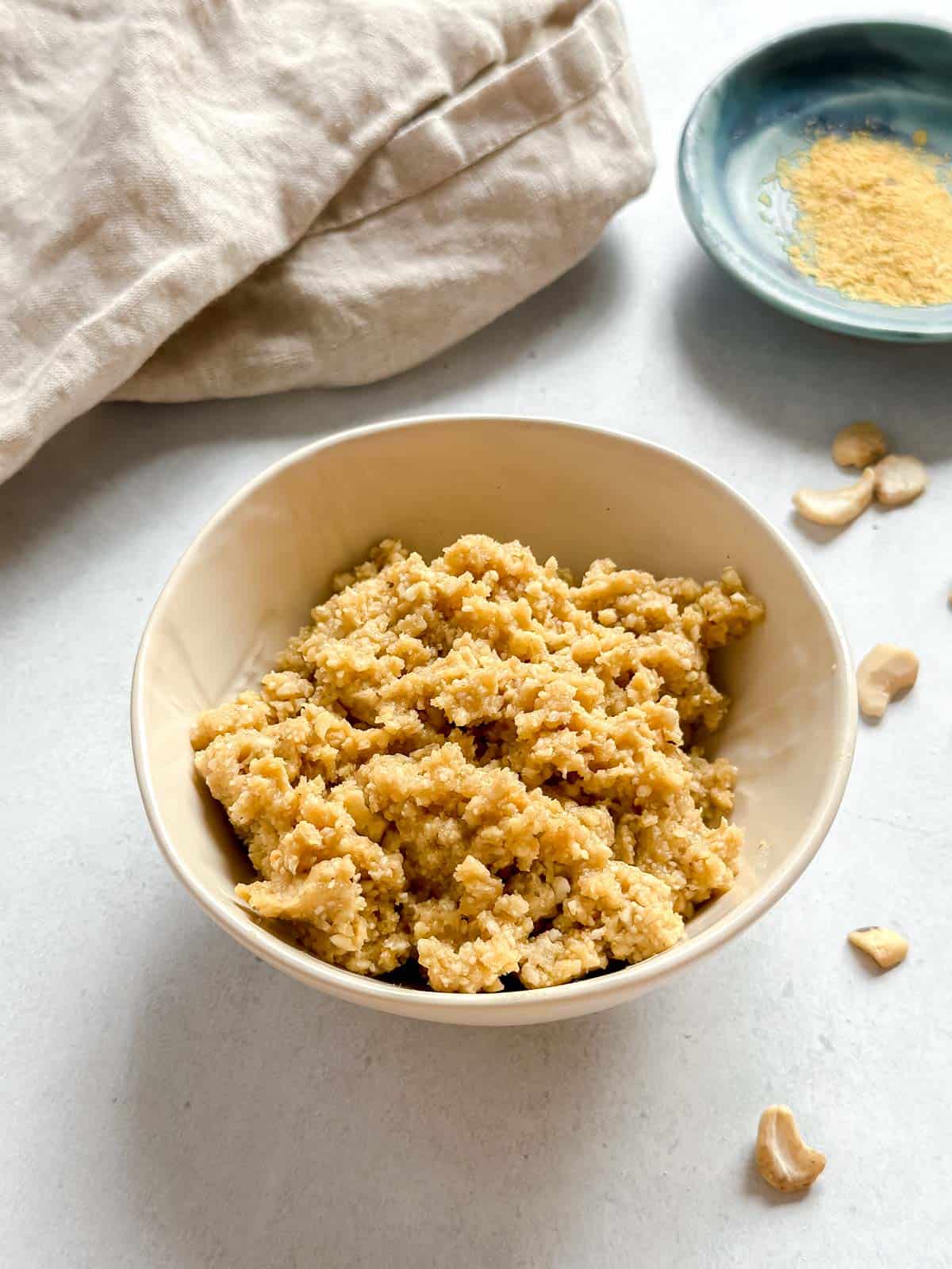 Crumbly vegan cotija cheese in bowl with loose cashews on white background. Nutritional yeast in small blue bowl and a tan napkin in background.