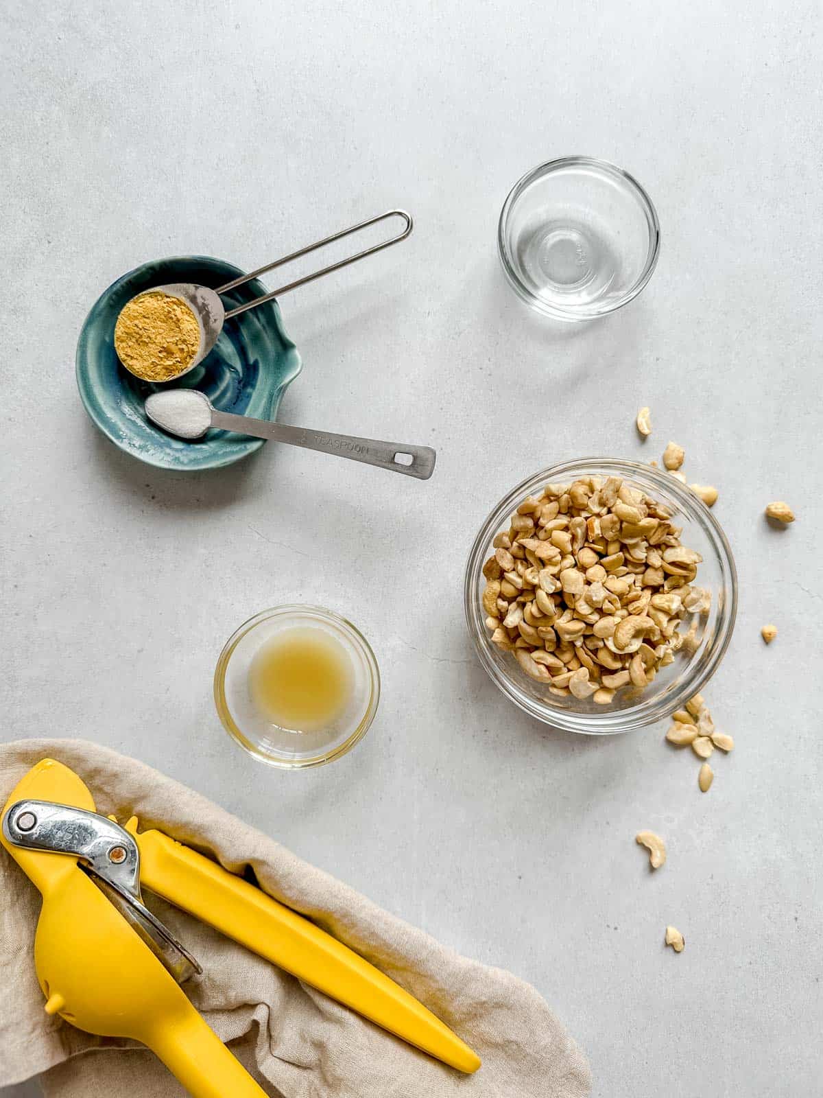 Individual ingredients placed in bowls: cashews, lemon juice, white vinegar along with nutritional yeast and salt in measuring spoons on a blue dish. Yellow lemon juicer on tan napkin. All on grey colored background.
