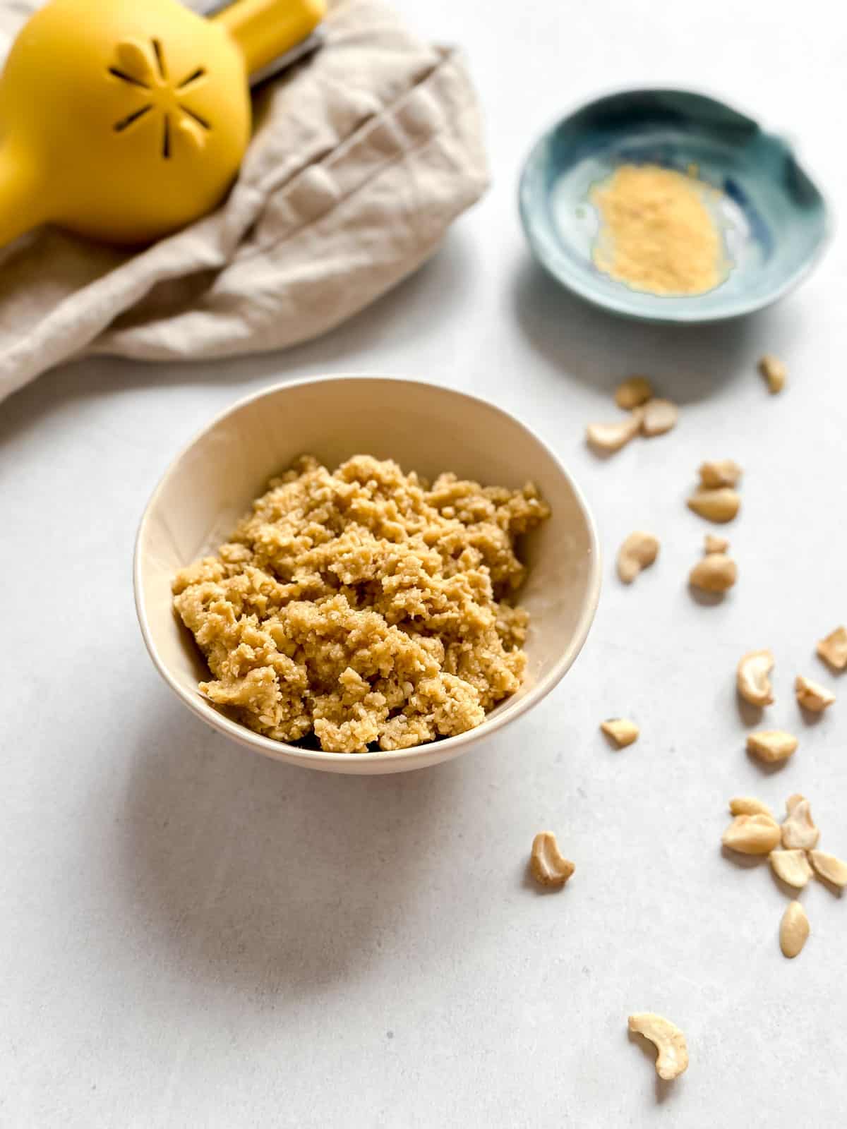 Crumbly vegan cotija cheese in bowl with loose cashews on white background. Nutritional yeast in small blue bowl and lemon juicer on tan napkin in background.