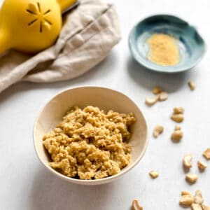 Crumbly vegan cotija cheese in bowl with loose cashews on white background. Nutritional yeast in small blue bowl and lemon juicer in background.