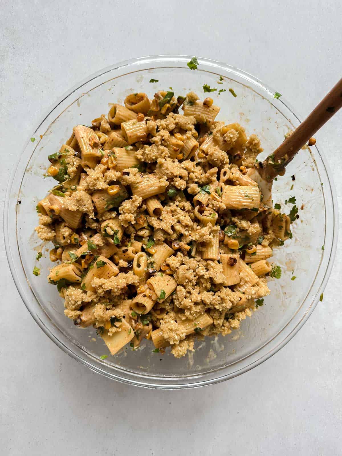Glass bowl filled with mixed street corn pasta salad topped with vegan cotija cheese and a wooden spoon