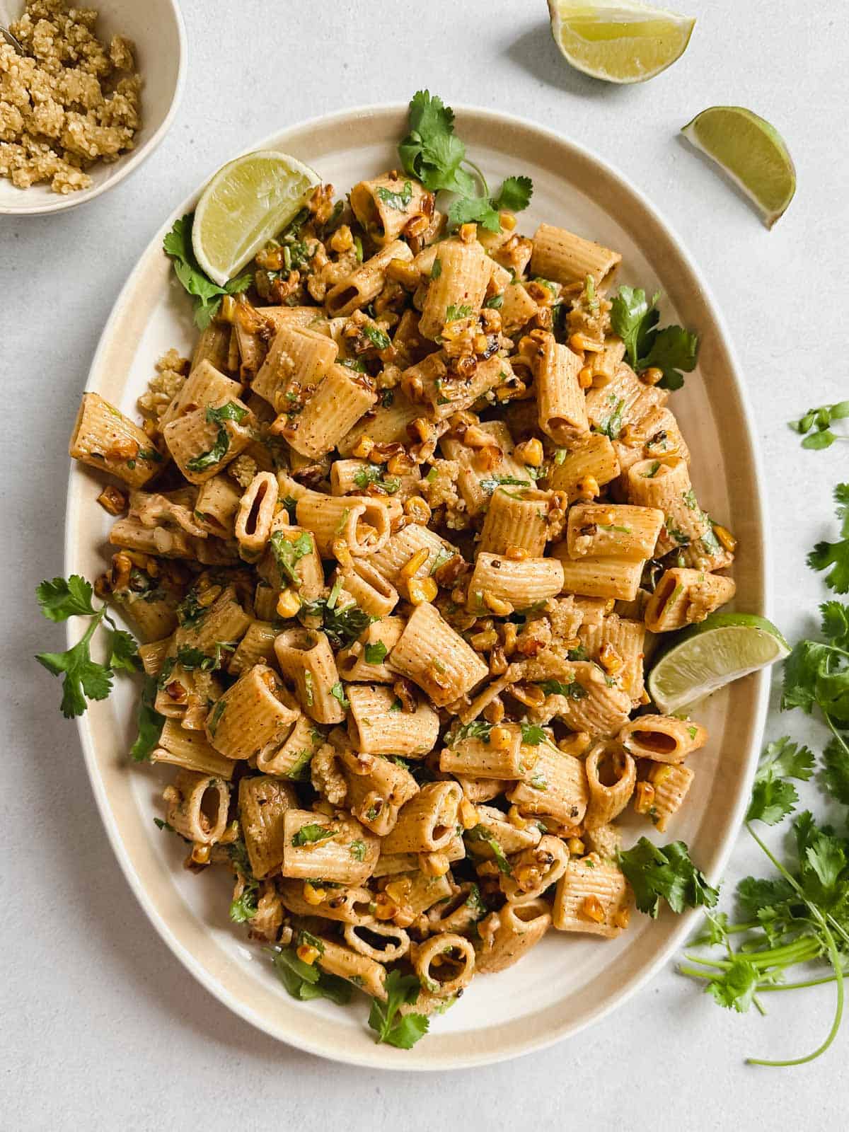 Vegan street corn pasta salad on an oval serving dish garnished with lime, cilantro, cotija cheese on a white background.