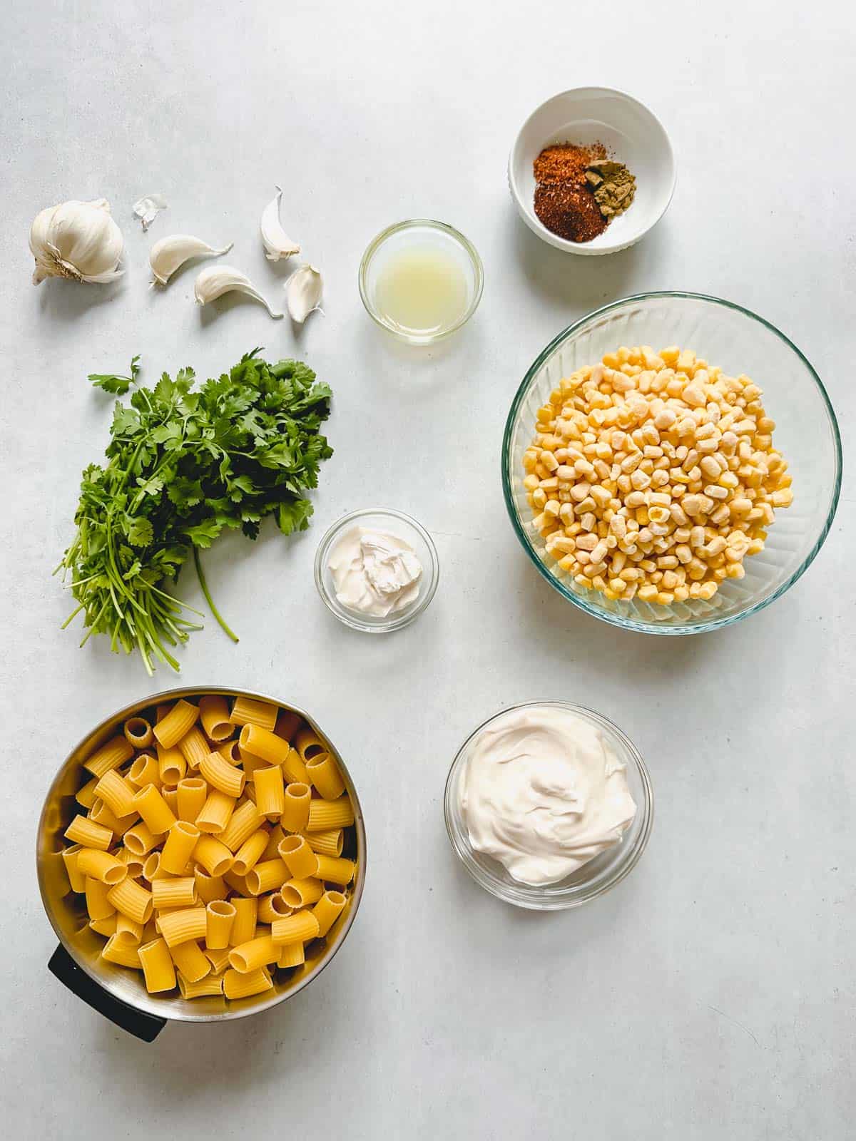 Ingredients in individual bowls: uncooked pasta, vegan mayonnaise, vegan sour cream, frozen corn, lime juice, spices, fresh cilantro and garlic gloves.