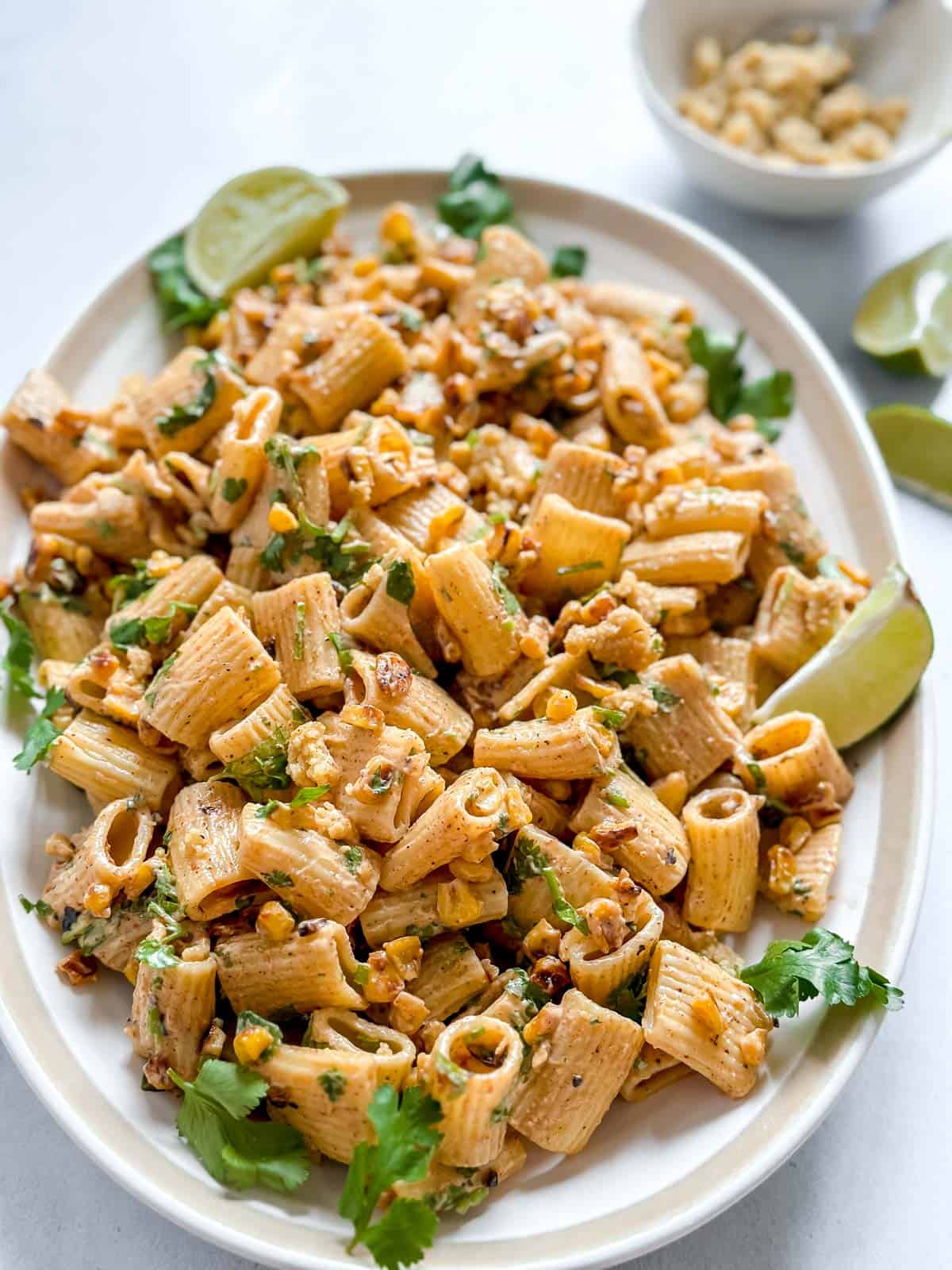 Vegan street corn pasta salad served on platter on a white back ground. Pasta is garnished with cilantro and lime wedges
