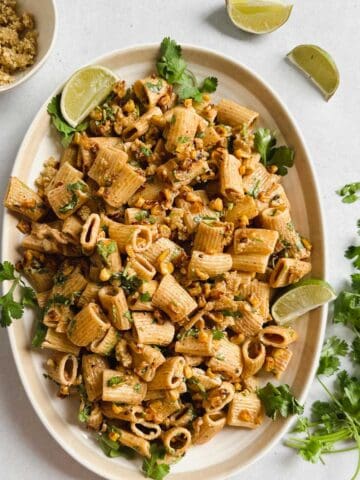 Vegan street corn pasta salad served on oval dish garnished with fresh cilantro, limes, and vegan cotija cheese.