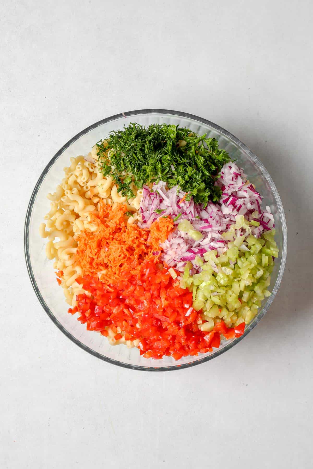 Cooked noodles, chopped vegetables, and herbs in large glass mixing bowl on a grey background
