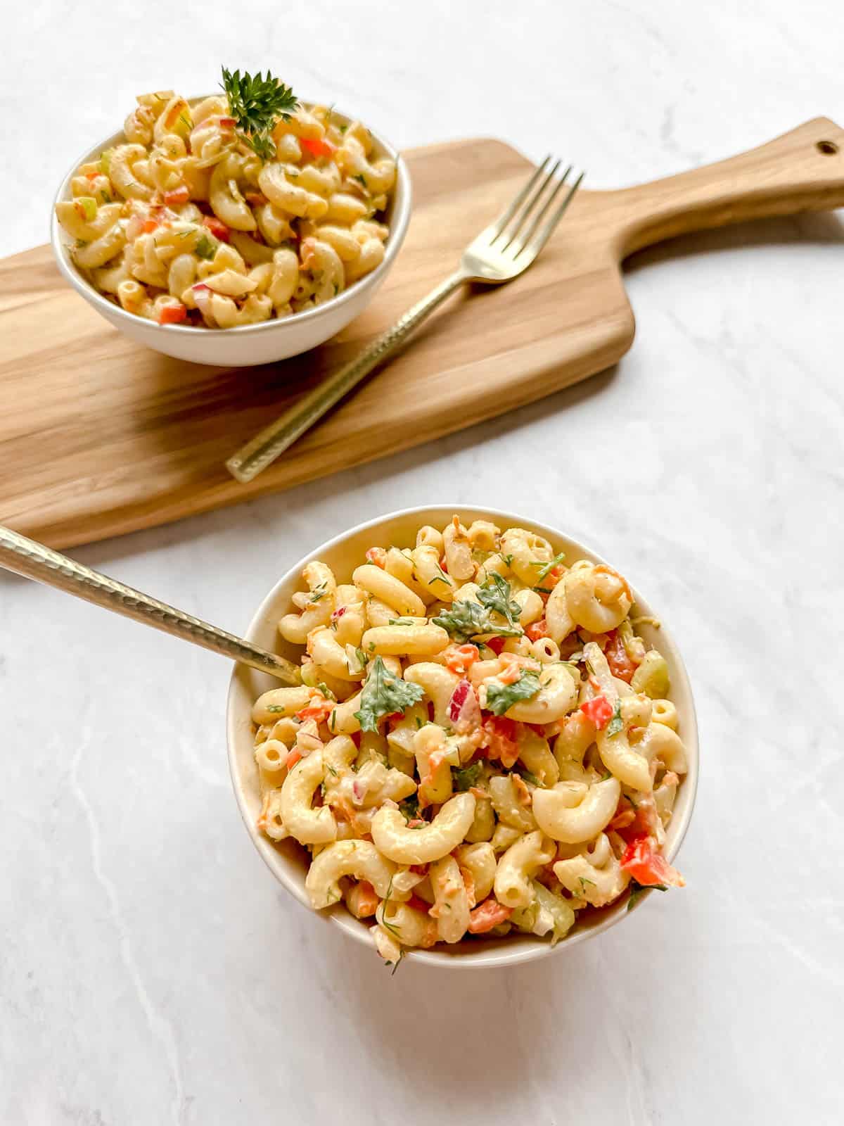 Macaroni salad in two white bowls along with 2 golden forks. One bowl is on a marble background and the other is on a wood platter
