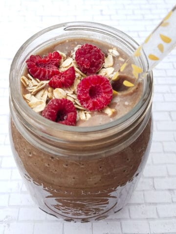 Chocolate smoothie in glass jar topped with raspberries and oats with a straw on a white background