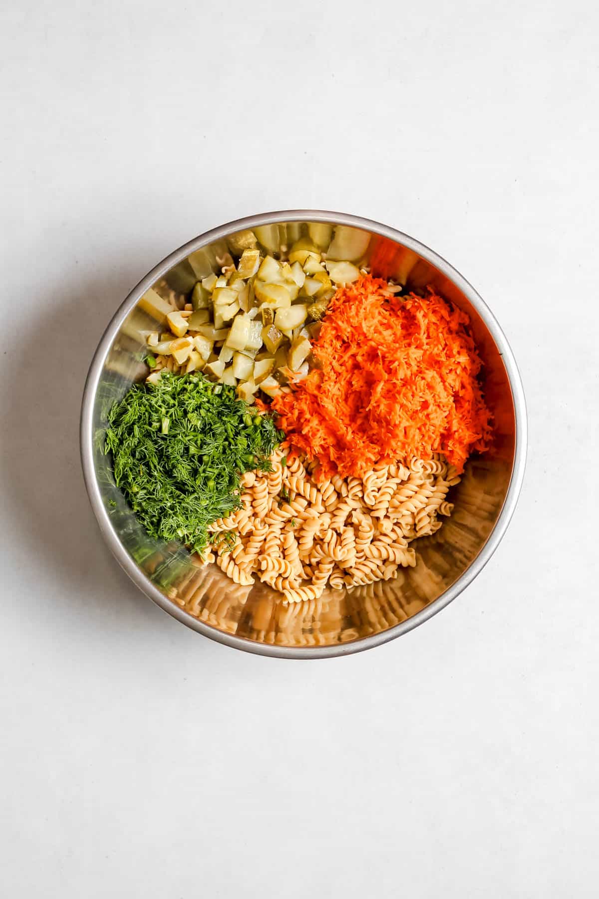 Cooked pasta, shredded carrot, chopped pickles and fresh dill in stainless steel mixing bowl